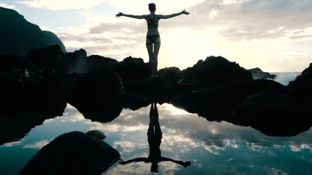 日落时分，一个女人在空中举起双臂，观察巨大的海浪冲击岩石并喷洒。电影慢动作. — 图库视频影像