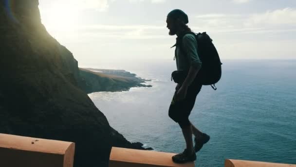 Silhouette de jeune randonneur barbu confiant avec sac à dos marchant sur le bord d'une route dans les îles Canaries au-dessus de l'océan . — Video