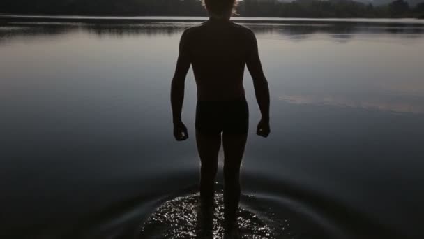 Backside cara vista caminha para a água de um lago de montanha e nada longe ao nascer do sol. A silhueta de homem contra reflexões belas de luz solar em água de um lago montês . — Vídeo de Stock