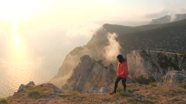 Jonge man loopt langs de rand van een klif observeren prachtige dramatische kleurrijke zonsondergang boven een zee van een hoge berg in de Krim. — Stockvideo