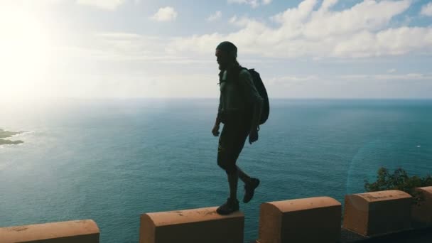 Silhouette eines jungen selbstbewussten bärtigen Wanderers mit Rucksack, der am Rande einer Straße auf Kanarischen Inseln hoch über dem Ozean spaziert. — Stockvideo