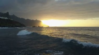 Ufukta güzel gün batımı ve dağlara bakan okyanus dalgaları üzerinde uçuş. Tenerife, Kanarya Adaları, İspanya'da güzel gün batımı havadan drone atış