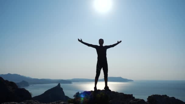 若い長い髪の男は、美しい青空に対して海の上の山の頂上に立って手を上げます。頂上に立つ幸せなハイカー男のシルエット. — ストック動画