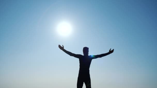 若い長い髪の男は、美しい青空に対して海の上の山の頂上に立って手を上げます。頂上に立つ幸せなハイカー男のシルエット. — ストック動画