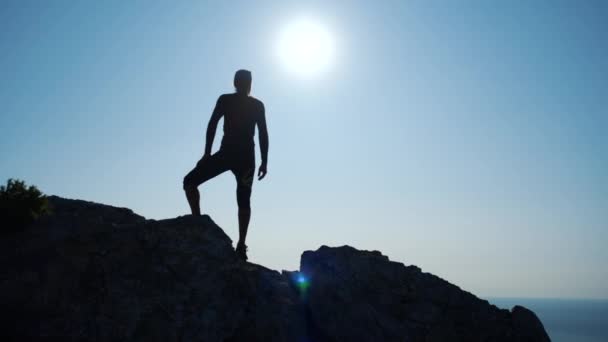Ο νεαρός εμπνευσμένος άνθρωπος στέκεται στην κορυφή ενός βουνού πάνω από τη θάλασσα ενάντια στον όμορφο γαλάζιο ουρανό. Σιλουέτα ενός ευτυχισμένο πεζοπόρο που στέκεται στη σύνοδο. — Αρχείο Βίντεο
