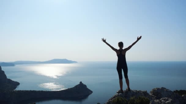 Mujer joven inspirada levanta las manos de pie en la cima de una montaña sobre el mar contra el hermoso cielo azul. Silueta de una mujer excursionista feliz de pie en la cumbre . — Vídeos de Stock
