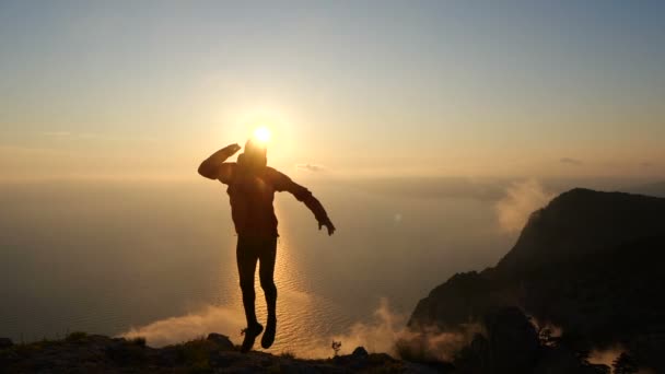 スローモーションで美しい夕日に対して空中でジャンプし、足をつかむ楽観的な若者のシルエット. — ストック動画