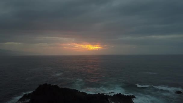 Vol lent vers l'arrière au-dessus de l'océan orageux vers un beau coucher de soleil coloré. Drone aérien de vagues océaniques roulant sur des roches noires . — Video