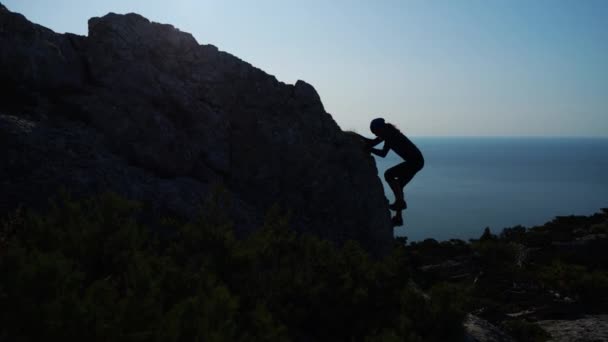 Ung långhårig inspirerad man höjer sina händer upp stående på toppen av ett berg ovanför havet mot vackra blå himmel. Silhuett av en lycklig vandrare man står på toppmötet. — Stockvideo