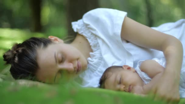母亲亲吻睡在户外绿草下的婴儿。绿色夏日公园里快乐的年轻母子。美丽的家庭在春天公园享受大自然. — 图库视频影像