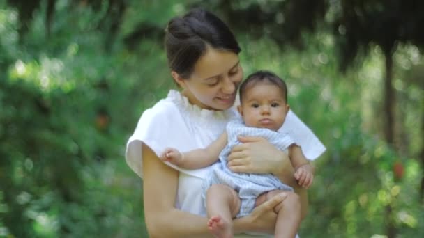 Mamma kysser sitt barn hålla barnet i händerna på en grön bakgrund av gräs och träd. Lycklig ung mamma håller sitt barn i Green Park. Vacker familj i Spring Park njuter av naturen utomhus. — Stockvideo
