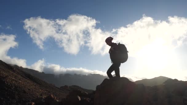 Silhouet van een jonge vrouw wandelaar met rugzak wandelen naar de top tegen blauwe hemel en wolken. Slow Motion. Lady is wandelen in prachtige bergen op Canarische eilanden. — Stockvideo