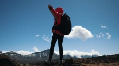 Siyah sırt çantası kolları uzanmış genç kadın yürüyüşçü mavi gökyüzü ve bulutlara karşı Dağlar'da yüksek zirvede kalır. Yavaş çekim. Lady hiker Kanarya Adaları güzel dağ manzarası sahiptir.