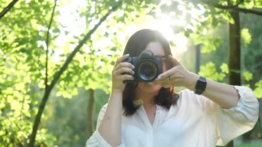 Beyaz gömlek giyen Güzel gülümseyen kız fotoğrafçı yeşil yaprakları yumuşak bir arka plan üzerinde bir parkta fotoğraf yapıyor.