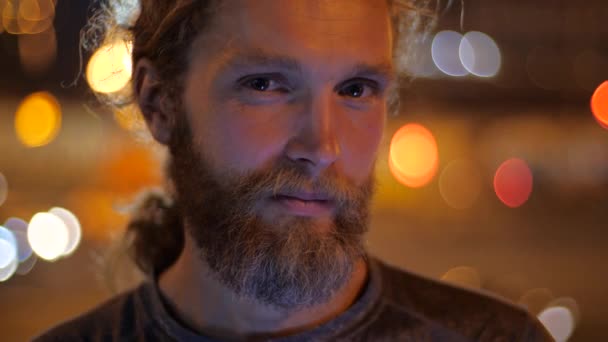 Close up, Portrait of a handsome Caucasian bearded long-haired young man looking straight to the camera and smiling on the background of moving evening lights of a city. — Stock Video