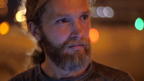 Gros plan, Portrait d'un beau jeune homme barbu caucasien aux cheveux longs regardant droit et souriant sur le fond des lumières du soir mouvantes d'une ville . — Video