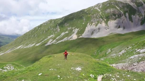 Вид с воздуха на молодую женщину с рюкзаком и пешеходными палками на горном маршруте среди альпийских лугов и скалистых скал Адыгеи. Удивительный горный пейзаж, красивые облака, покрывающие высокие горы . — стоковое видео