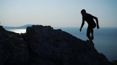 Genç uzun saçlı ilham adam güzel mavi gökyüzüne karşı denizüzerinde bir dağın tepesinde ayakta ellerini kaldırır. Zirvede duran mutlu bir yürüyüşçü adamın silueti.