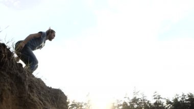 Genç uzun saçlı adam gün batımında açık havada yavaş çekim bir uçurumdan bir kum atlar.