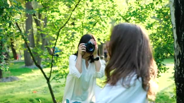 Menina bonita fotógrafo profissional vestindo camisa branca está fazendo fotos de uma menina sorridente feliz em um parque em um fundo macio de folhagem verde e água pulverizadora . — Vídeo de Stock