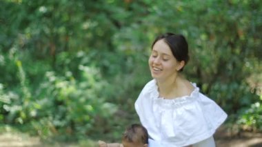 Genç Pretty Anne Yavaş Hareket bir parkta yeşillik yeşillik yeşil bir arka plan üzerinde onu yeni doğan kızı yetiştirme. Mutlu genç Anne ve Çocuk açık havada oynamak ve doğanın tadını çıkarın.