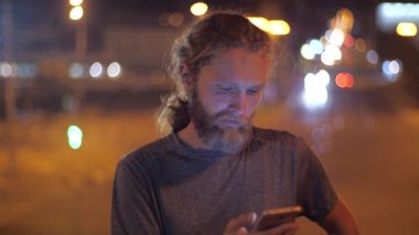Yakın çekim, Bir şehrin hareketli akşam ışıkları arka planda akıllı telefon kullanarak yakışıklı bir Kafkas sakallı uzun saçlı genç adam Portresi.