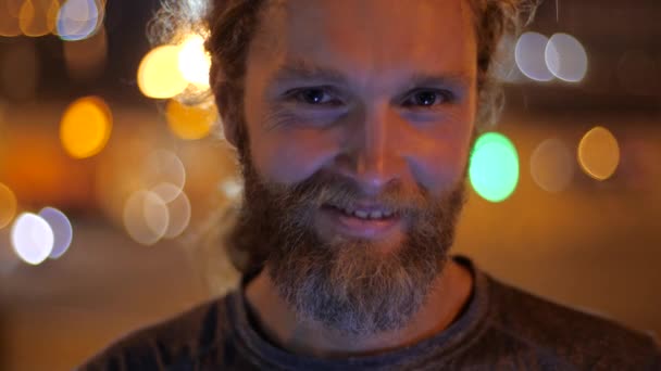 De cerca, Retrato de un guapo caucásico barbudo de pelo largo joven mirando directamente a la cámara y sonriendo en el fondo de las luces de la noche en movimiento de una ciudad . — Vídeos de Stock