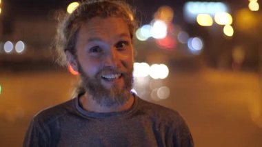 Gülümseyen yakışıklı Kafkas sakallı uzun saçlı genç adam kameraya doğru bakar, konuşur ve bir şehrin hareketli akşam ışıkları arka planda başparmak gösterir.