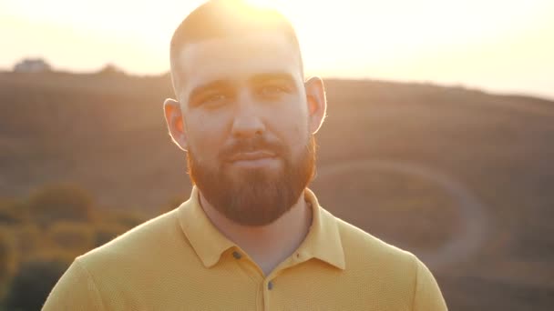 Close up, Retrato de um belo jovem caucasiano barbudo olhando diretamente para a câmera no fundo de um pôr do sol brilhante sobre a paisagem natural. Alegria da vida, estilo de vida ativo, blogueiro . — Vídeo de Stock