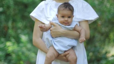 Genç güzel anne kucaklar, beşik ve Park yeşillik yeşil bir arka plan üzerinde onu yeni doğan kızı öper, yavaş hareket. Güzel küçük kız gülümseyen ve annelerin kollarında açık havada gülüyor.