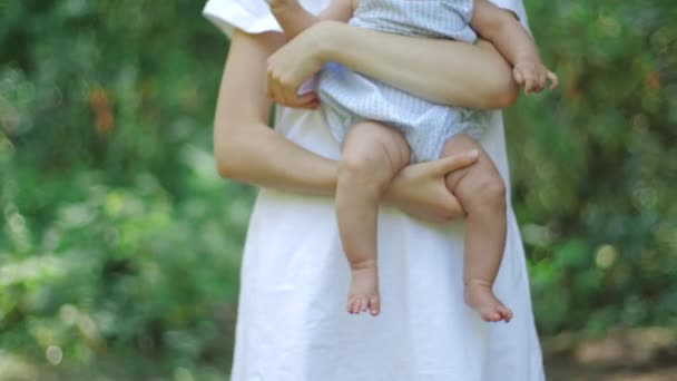 若い母親は、公園の葉の緑の背景に足で彼女の生まれたばかりの娘を抱きしめ、抱きしめ、揺りかご, スローモーション.屋外でママの腕に座って美しい小さなスワーシーな女の子. — ストック動画