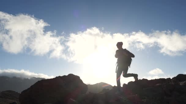 Silhuett av en ung kvinna vandrare med ryggsäck går uppför mot toppmötet mot blå himmel och moln vid solnedgången. Slow motion. Lady är vandring i vackra berg på Kanarieöarna. — Stockvideo