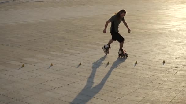 Jonge langharige man rolschaatser danst tussen kegels in een leuke avond in een stadspark. Freestyle slalom Rolschaatsen tussen kegels in slow motion. — Stockvideo