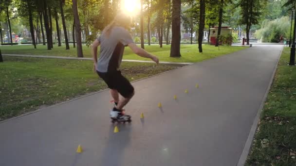 Young long-haired bearded man roller skater is dancing between cones in a nice evening in a city park. Freestyle slalom Roller skating between cones in slow motion. — Stock Video
