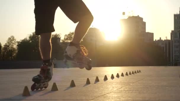 Ein junger langhaariger Mann läuft an einem schönen Abend im Stadtpark zwischen Zapfen Schlittschuh. Freestyle Slalom Rollschuhfahren zwischen Kegeln in Zeitlupe. — Stockvideo