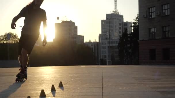 Jovem homem de cabelos longos está patinando profissionalmente entre cones em um pôr do sol agradável noite em um parque da cidade. slalom Freestyle Patinação de rolo entre cones . — Vídeo de Stock