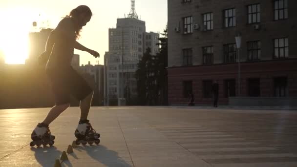 Joven hombre de pelo largo es patinaje profesional entre conos en una bonita puesta de sol por la noche en un parque de la ciudad. Freestyle slalom Patinaje sobre ruedas entre conos . — Vídeo de stock