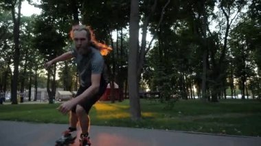 Genç uzun saçlı sakallı adam patenci başparmak gösteren bir şehir parkında güzel bir akşam dans ediyor. Gün batımında Serbest Stil Paten.