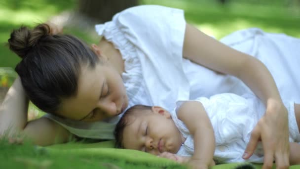 母亲亲吻睡在户外绿草下的婴儿。快乐微笑的年轻母子在绿色的夏天公园。美丽的家庭在春天公园享受大自然. — 图库视频影像