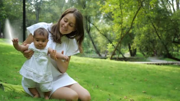 Jeune jolie mère en robe blanche apprend à sa fille à marcher sur une herbe verte fraîche dans un parc. Joyeux jeune mère et enfant apprenant à marcher à l'extérieur et profiter de la nature . — Video