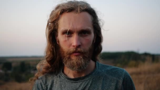 De cerca, Retrato de un guapo joven caucásico barbudo con el pelo largo que fluye mirando amablemente a la cámara en el fondo de una puesta de sol brillante sobre la naturaleza. Alegría de la vida, estilo de vida activo . — Vídeo de stock