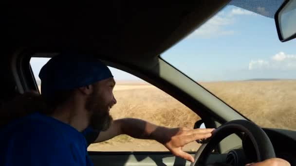 Homem barbudo feliz dirige um carro na paisagem rural. Motorista ondas mão inclinada para fora da janela lateral — Vídeo de Stock