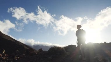 Sırt çantası mavi gökyüzü ve bulutlara karşı zirveye doğru yürüyen genç bir kadın yürüyüşçü silueti. Yavaş çekim. Lady Kanarya Adaları güzel dağlarda yürüyüş.