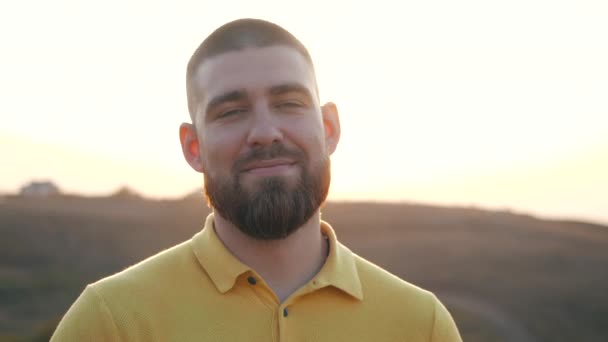 Da vicino, Ritratto di un bel giovane caucasico barbuto che guarda la macchina fotografica e di lato, sorridendo e mostrando i denti sullo sfondo di un tramonto sulla natura. Gioia della vita, stile di vita attivo . — Video Stock