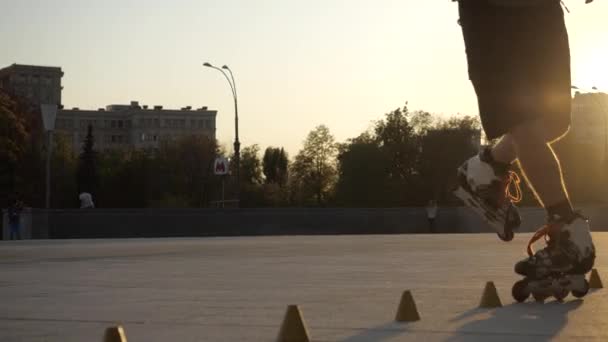 Jonge langharige man rolschaatser danst 's avonds tussen kegels op een stadsplein bij zonsondergang. Freestyle slalom Rolschaatsen tussen kegels in slow motion. — Stockvideo