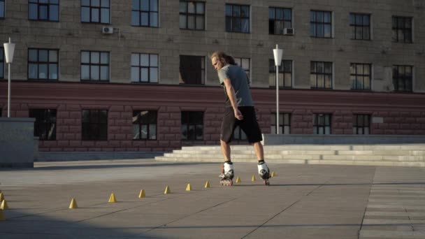 Genç, uzun saçlı kaykaycı adam akşamleyin şehir meydanında koniler arasında dans ediyor. Serbest stil slalom patenler ağır çekimde koniler arasında kayıyor. — Stok video