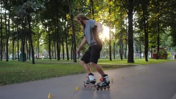 Mladý vousatý krasobruslař tančí mezi kužele v hezkém večeru v městském parku. Freestyle slalom kolečkové bruslení mezi kužele v pomalém pohybu. — Stock video