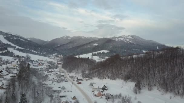 Vlucht over een dorp in de Karpaten en een ski-oord ernaast. Birds Eye uitzicht op besneeuwde huizen in bergen. Landelijk landschap in de winter. Karpaten dorp in de sneeuw van een hoogte. — Stockvideo