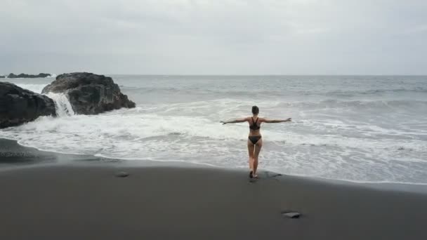 スペイン、テネリフェ島の黒い砂浜で水に流れ込む女性の航空写真。海の波が壊れ、黒い砂の上に白い泡を作成します, カナリア諸島. — ストック動画