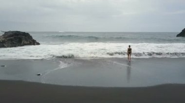 İspanya'nın Tenerife'deki siyah kumlu plajında suya giren ve yükselen elleri yükselen güzel bir kadının havadan görünümü. Okyanus dalgaları kırmak ve siyah bir kum üzerinde beyaz köpük oluşturmak, Kanarya Adaları.